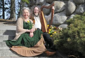 Kuvassa tarinankertoja Riikka Palonen ja muusikko Richard Raha-Aho. He istuvat kiviportailla kesäisessä maisemassa ja Richard pitelee sylissään harppua