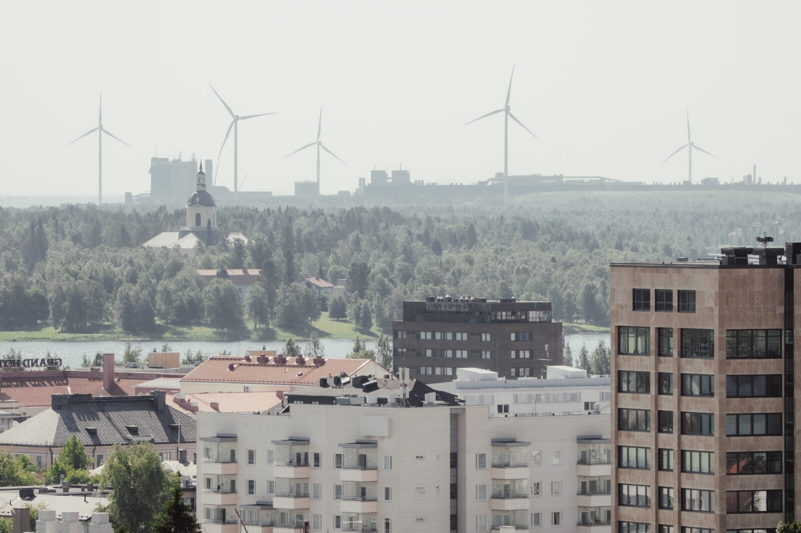 Kesäinen kaupunkimaisema, etualalla kerrostaloja, taustalla horisontissa tuulimyllyjä.