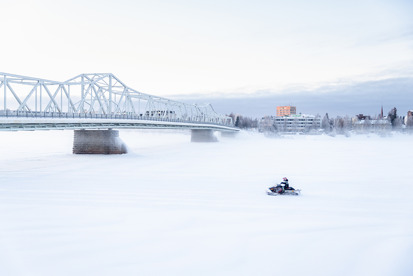 Moottorikelkkailija ajaa joen jäällä lumisessa maisemassa, taustalla kaupungin keskustan korkeita taloja.